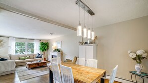 A spacious living area with a wooden dining table, white chairs, a sofa, and a large window. - Stroll to Shore! Michigan City Family Getaway (Michigan City)