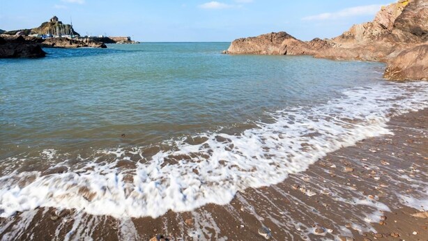 Ein Strand mit klarem Wasser, Felsformationen und einer weit entfernten Struktur.