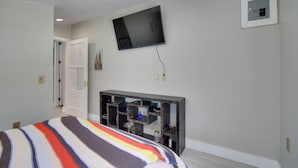A bedroom with a bed, a television mounted on the wall, a console table, and a door leading to another room. - Private Ocean Springs Waterfront Retreat w/Porch (Ocean Springs)
