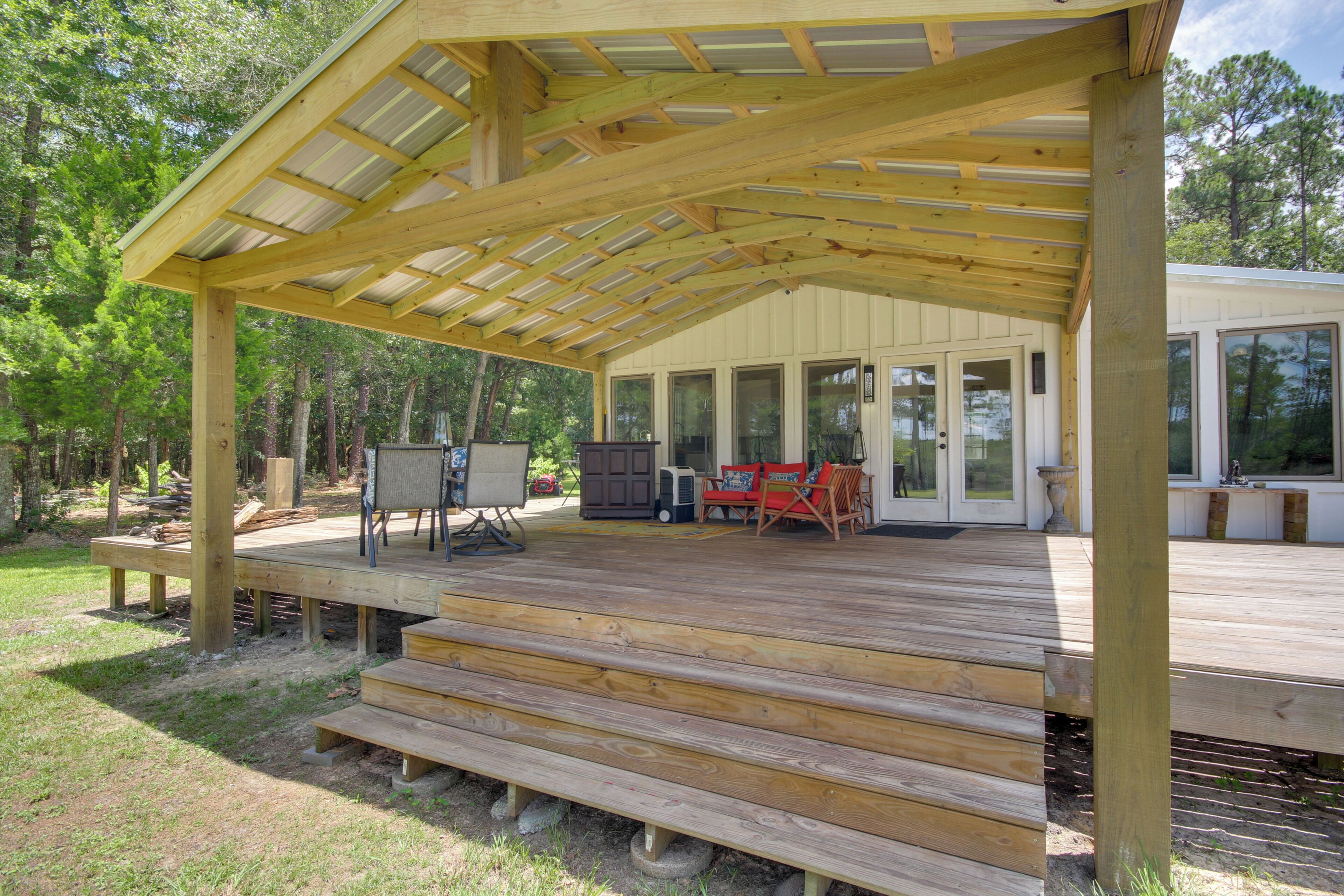 Une terrasse en bois couverte, offrant une vue sur une maison, du mobilier d’extérieur et un paysage forestier naturel en arrière-plan.