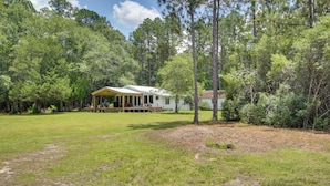 A single-story house with a covered porch, surrounded by a grassy yard and dense trees. - Private Ocean Springs Waterfront Retreat w/Porch (Ocean Springs)