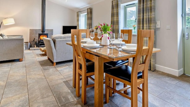 Un coin repas avec une table en bois dressée pour quatre personnes, un canapé, une cheminée et une vue sur l’extérieur.