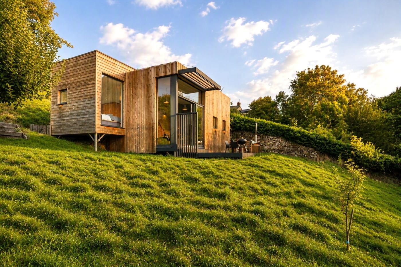 A modern wooden house with a deck, large windows, and a stone wall.