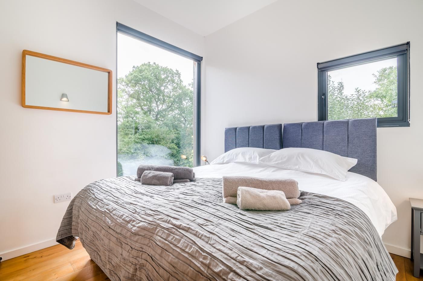 A modern bedroom with a large bed, two windows, and a view of greenery outside.