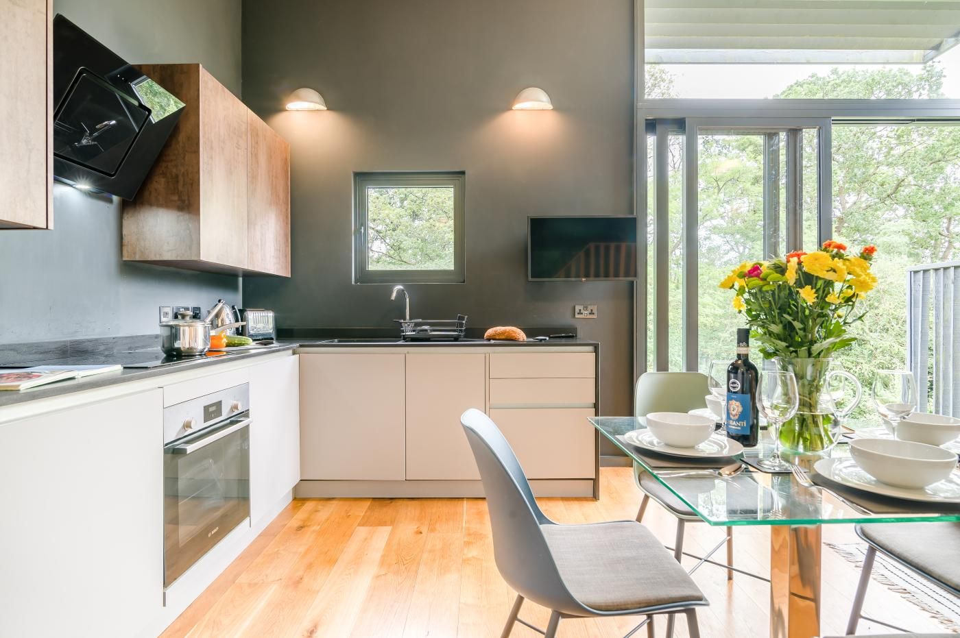 A modern kitchen with a dining area, wooden cabinets, a built-in oven, and a large window offering a view of greenery.
