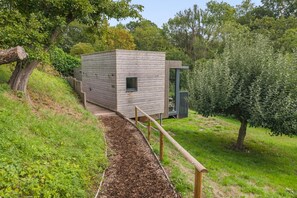 Eine moderne Holzhütte mit kleinem Fenster, umgeben von üppiger Vegetation und einer Holzterrasse, die darauf führt.