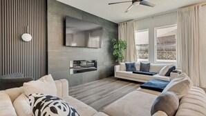 A modern living room with a sofa, a flat-screen TV, a ceiling fan, and a large window with curtains. - Great for Families! Glenn Heights Getaway w/ Yard (Glenn Heights)
