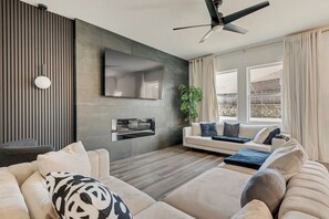 Ein modernes Wohnzimmer mit einer Couch, einem Flachbildfernseher, einem Deckenventilator und einem großen Fenster mit Vorhängen.