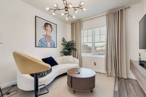 Ein modernes Wohnzimmer mit einer Couch, einem runden Couchtisch, einem Gemälde, einer Pflanze und einem großen Fenster mit Vorhängen.