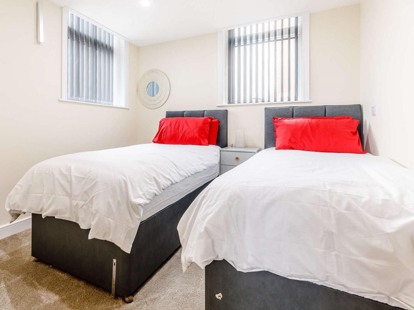 Habitación doble con dos camas, cada una con ropa blanca y almohadas rojas, una mesita de noche pequeña y un espejo redondo en la pared.