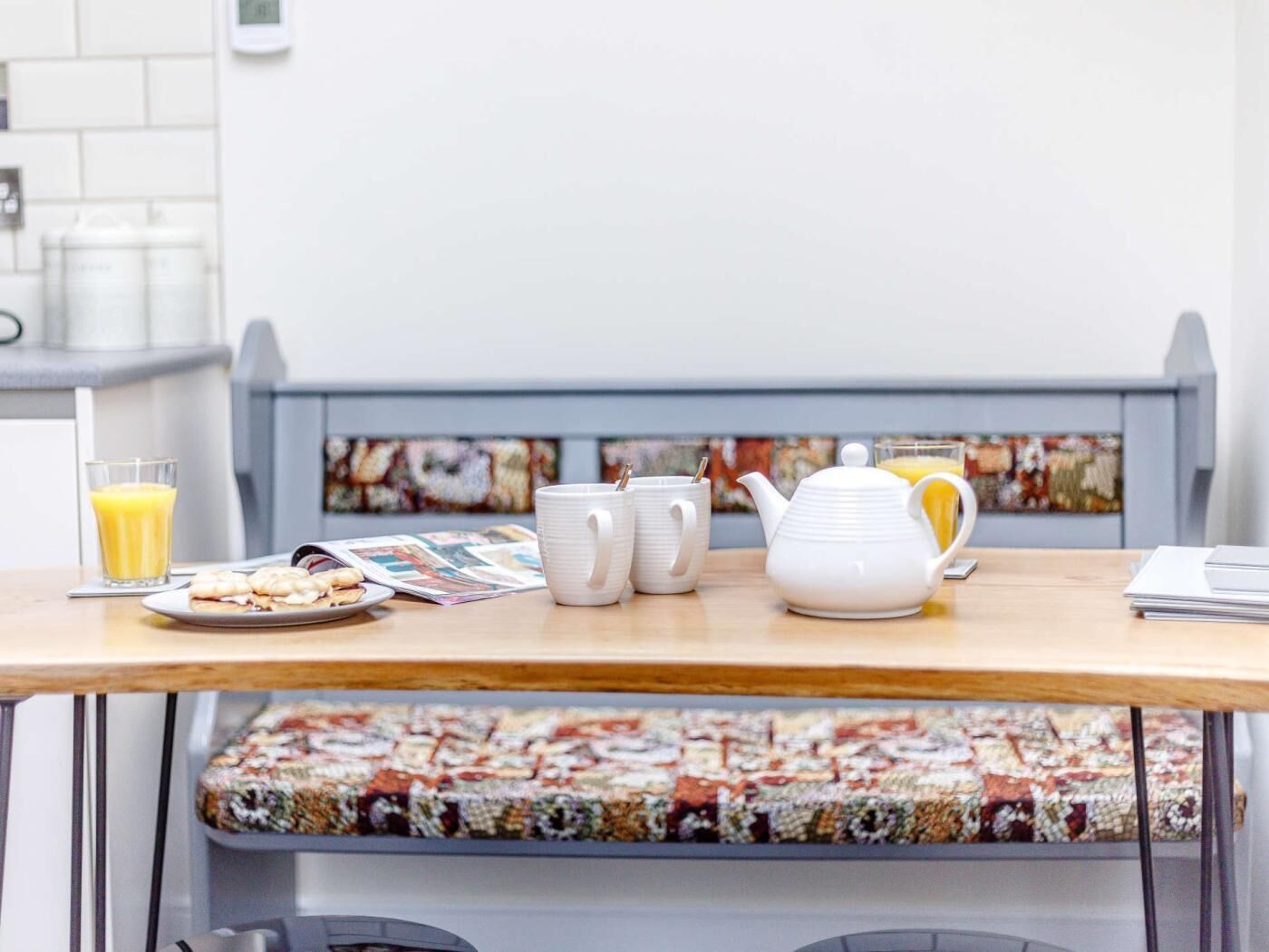 A breakfast table with a teapot, cups, a glass of orange juice, and a plate of cookies.