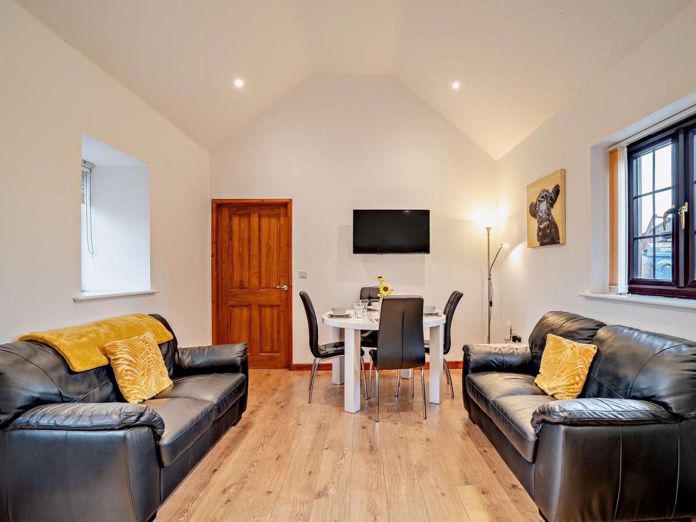 Un salon avec deux canapés en cuir noir, une table à manger avec des chaises, une porte en bois, un téléviseur fixé au mur et une fenêtre avec des rideaux.