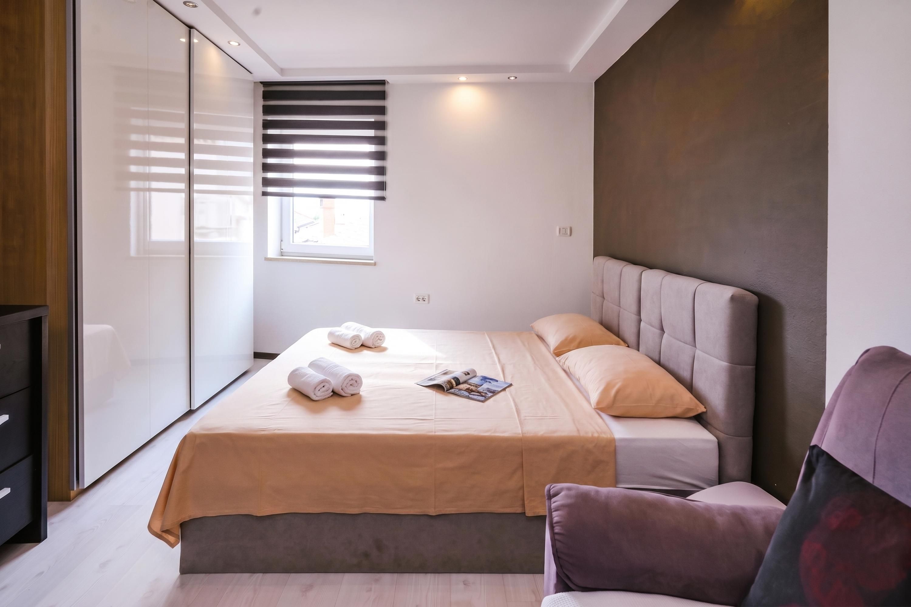 A modern bedroom with a bed, a chair, a window with blinds, and a wall-mounted TV.