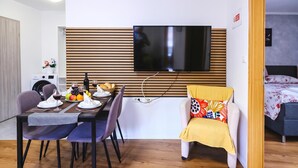 A modern living room with a dining area, a flat-screen TV mounted on a wooden panel wall, a sofa with a colorful cushion, and a bed with a floral bedspread. - Ota Residence Koper (Koper)
