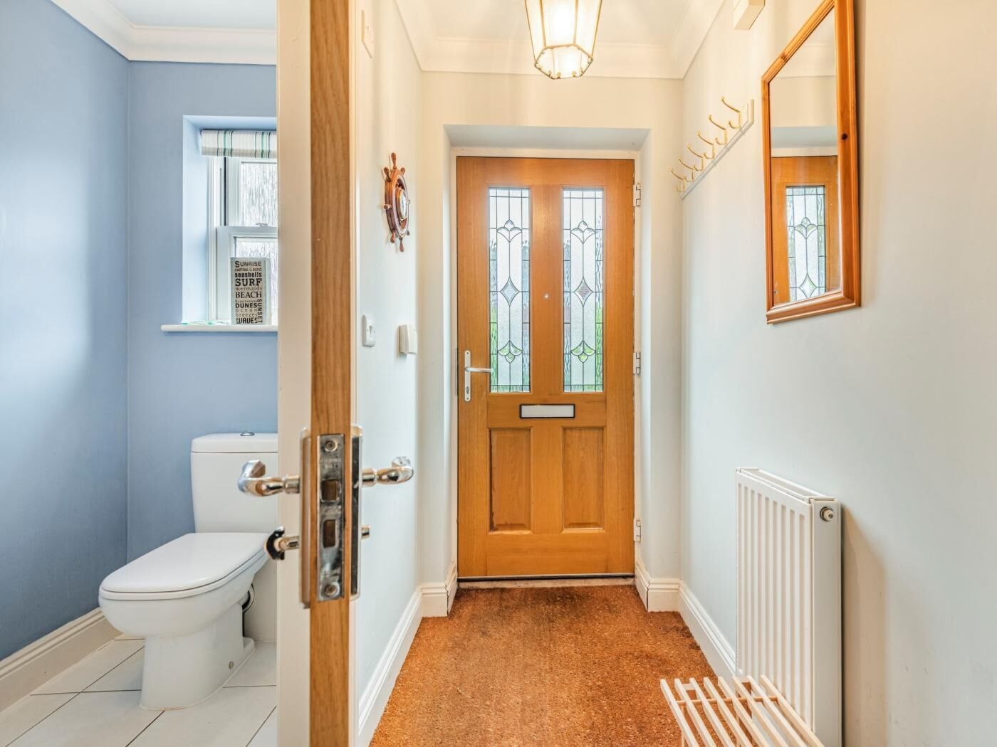 A hallway with a wooden door, a radiator, a white radiator, a white toilet, and a window with blinds.