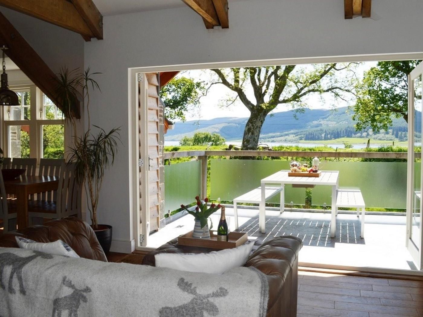 A cozy living room with a large window, a dining area with a table and chairs, and a view of a green landscape with a tree and mountains.