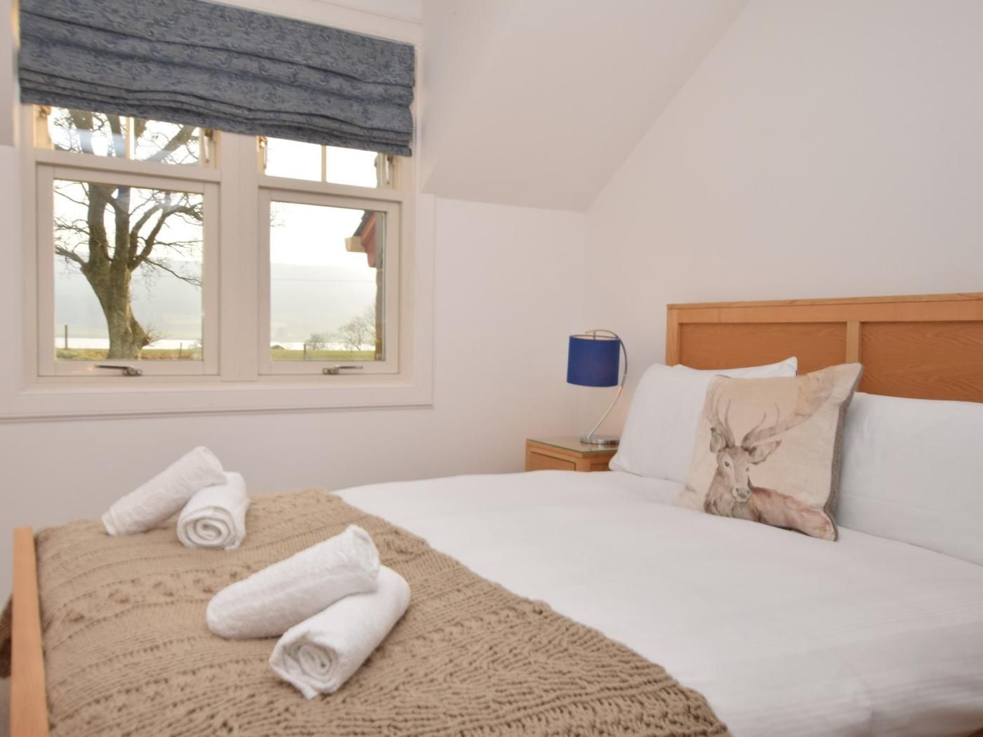 Ein Schlafzimmer mit Bett, Fenster mit Blick auf Bäume, Nachttisch mit Lampe und zusammengerollten Handtüchern auf dem Bett.