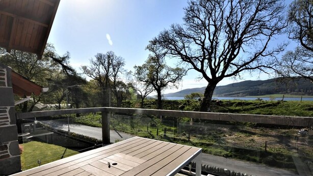 Um deck de madeira com guarda-corpo de vidro, com vista para uma área gramada, árvores e um corpo d'água ao fundo.