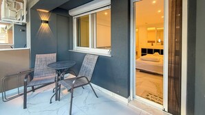 Un balcón con mesa y dos sillas, una ventana que da a un dormitorio y un aire acondicionado instalado en la pared.