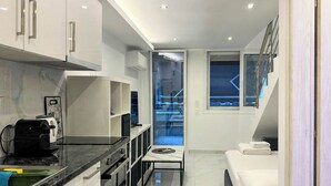 Una cocina moderna con cafetera, un área de comedor con mesa de vidrio, un sofá y vista al cielo azul por una ventana.