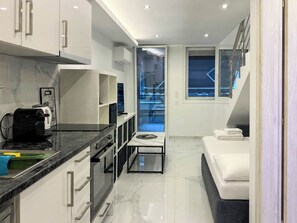 A modern kitchen with a coffee machine, a dining area with a glass table, a sofa, and a view of a blue sky through a window. - 23 - DIAS Suites by SDR (Paralia)