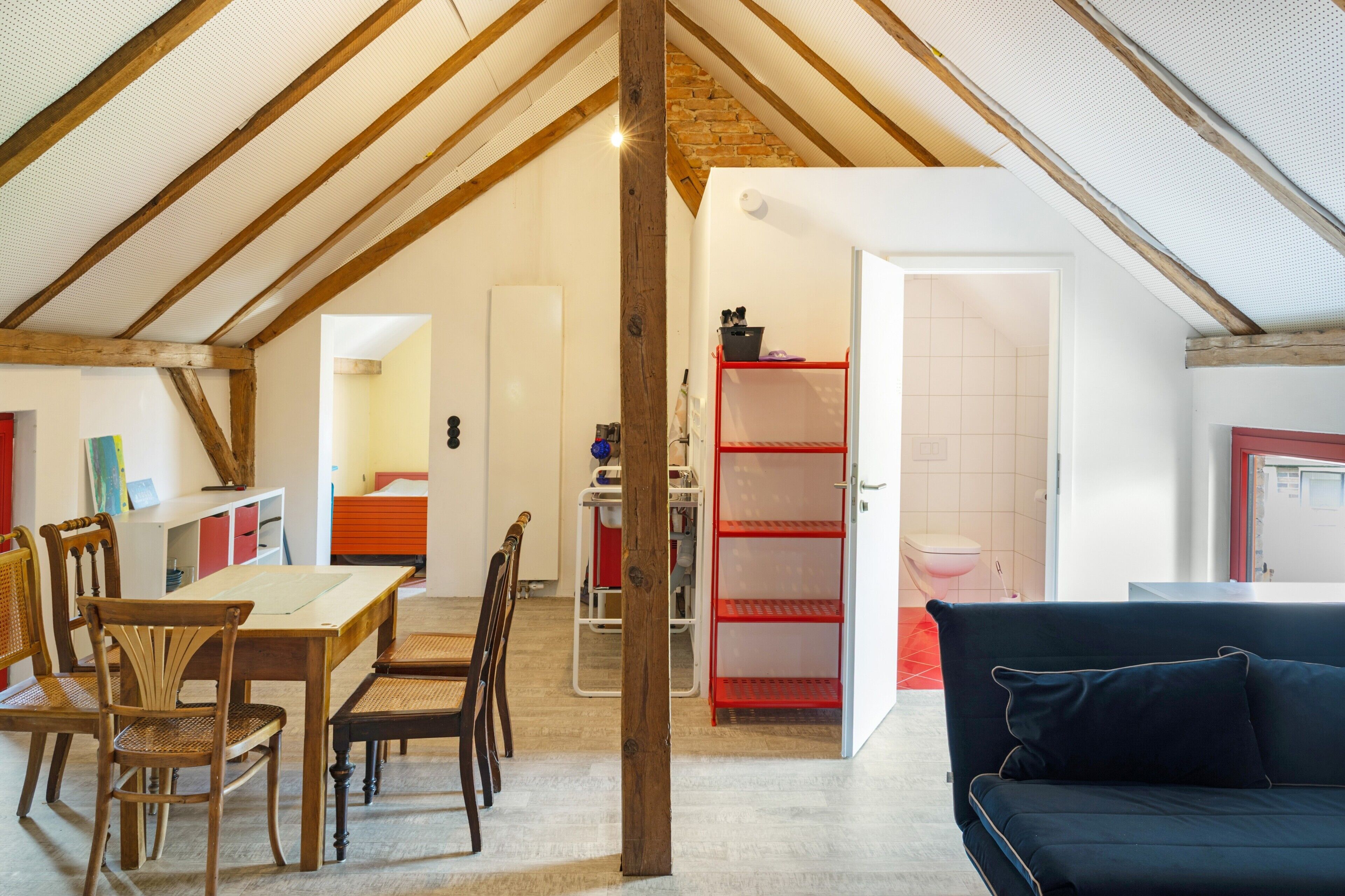 A spacious living area with a high ceiling, wooden beams, a dining table with chairs, a sofa, and a kitchen area with a red shelving unit.
