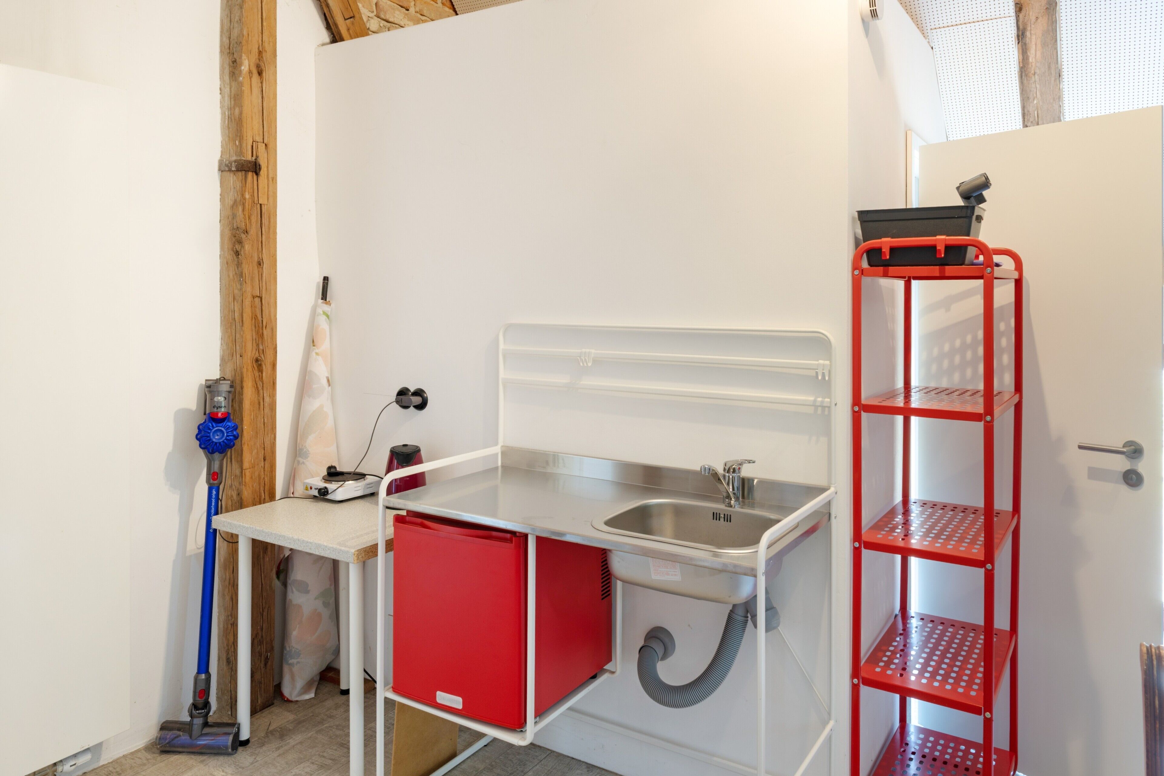 A compact kitchenette with a sink, a small storage unit, and a red metal shelf.