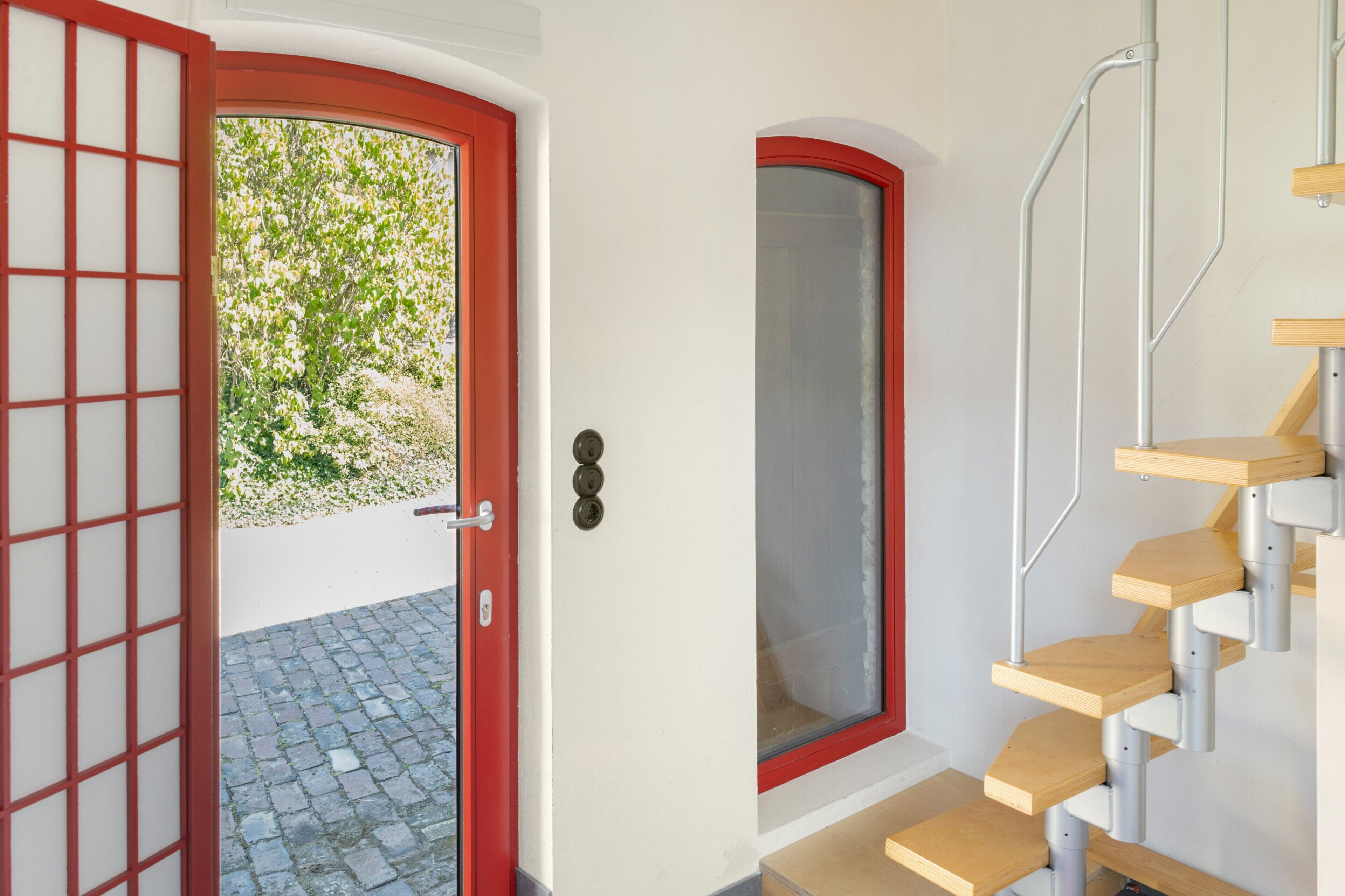 A red-framed door with a view of greenery outside, a white wall, and a wooden staircase with a metal handrail.
