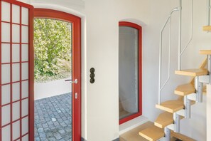 A red-framed door with a view of greenery outside, a white wall, and a wooden staircase with a metal handrail.