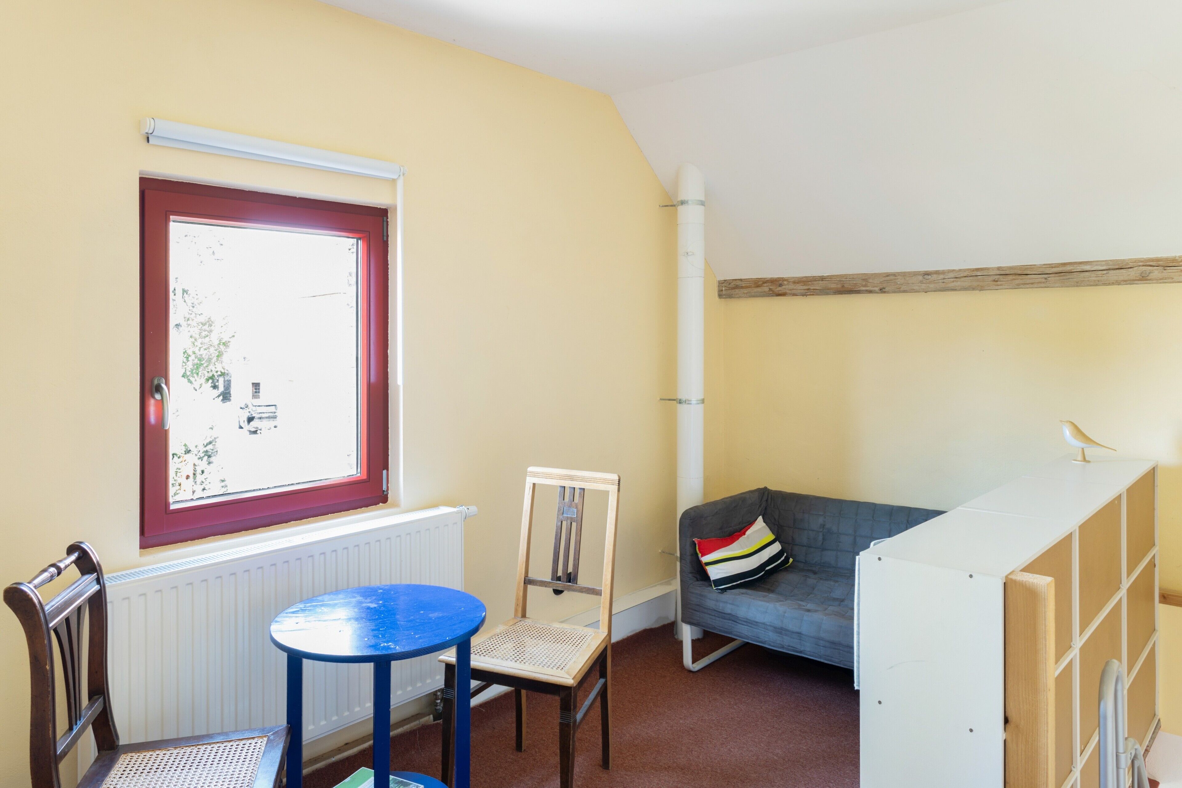 A room with a window, radiator, wooden chair, blue table, grey sofa, and a white cabinet.