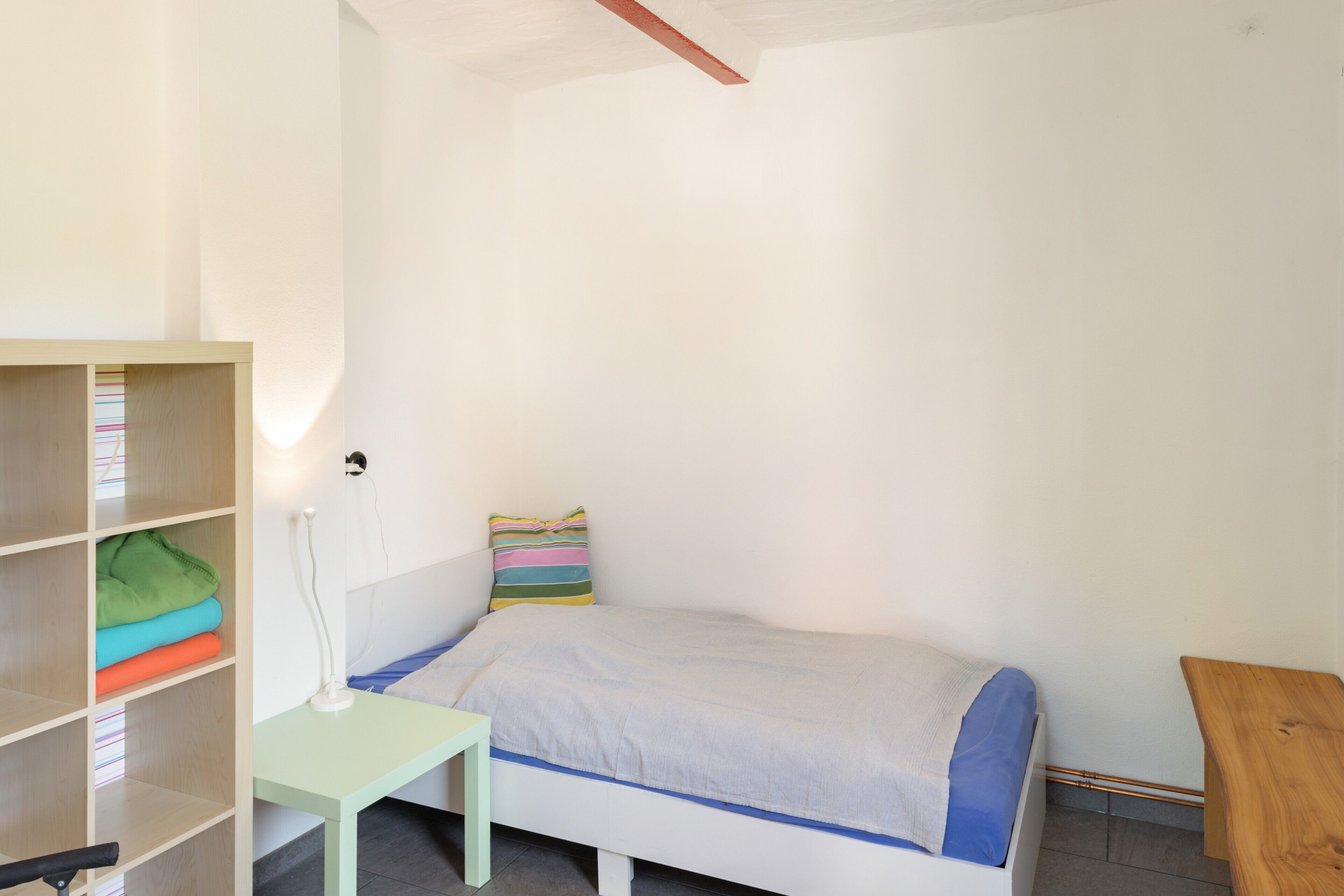 Una habitación pequeña con cama individual, pared blanca, estante de madera con ropa doblada y mesita de noche con lámpara.