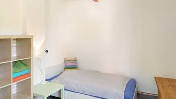 Una habitación pequeña con cama individual, pared blanca, estante de madera con ropa doblada y mesita de noche con lámpara.