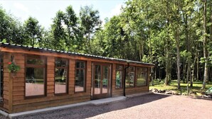 En træhytte med store vinduer, en grusvej og en skov i baggrunden.