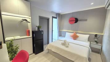 Moderni hotellihuone, jossa on sänky, työpöytä, tuoli, jääkaappi ja seinälle kiinnitetty kello.