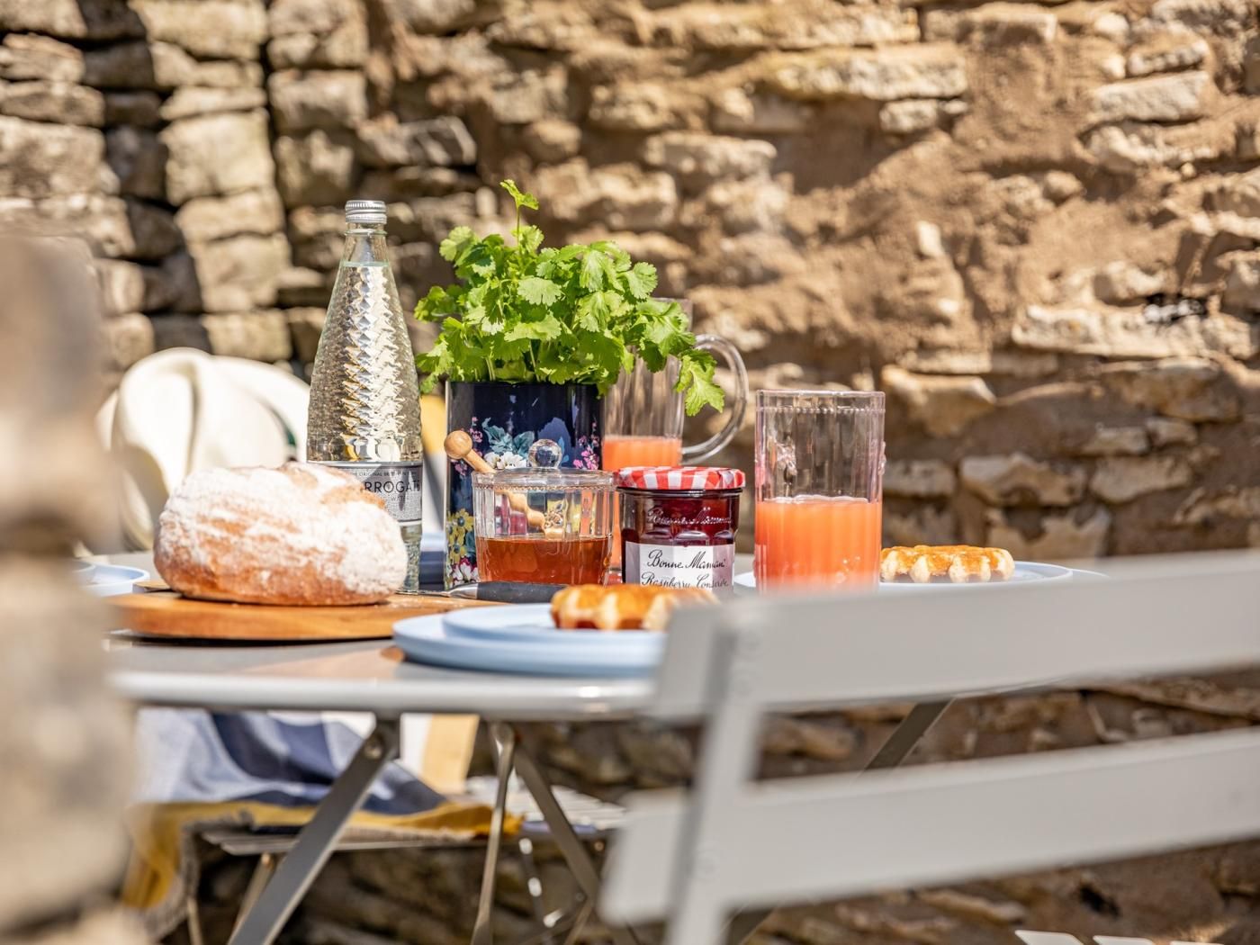 A table set outdoors with a stone wall background, featuring a bottle, a glass jar, a glass pitcher, a loaf of bread, and a plate with a pastry.