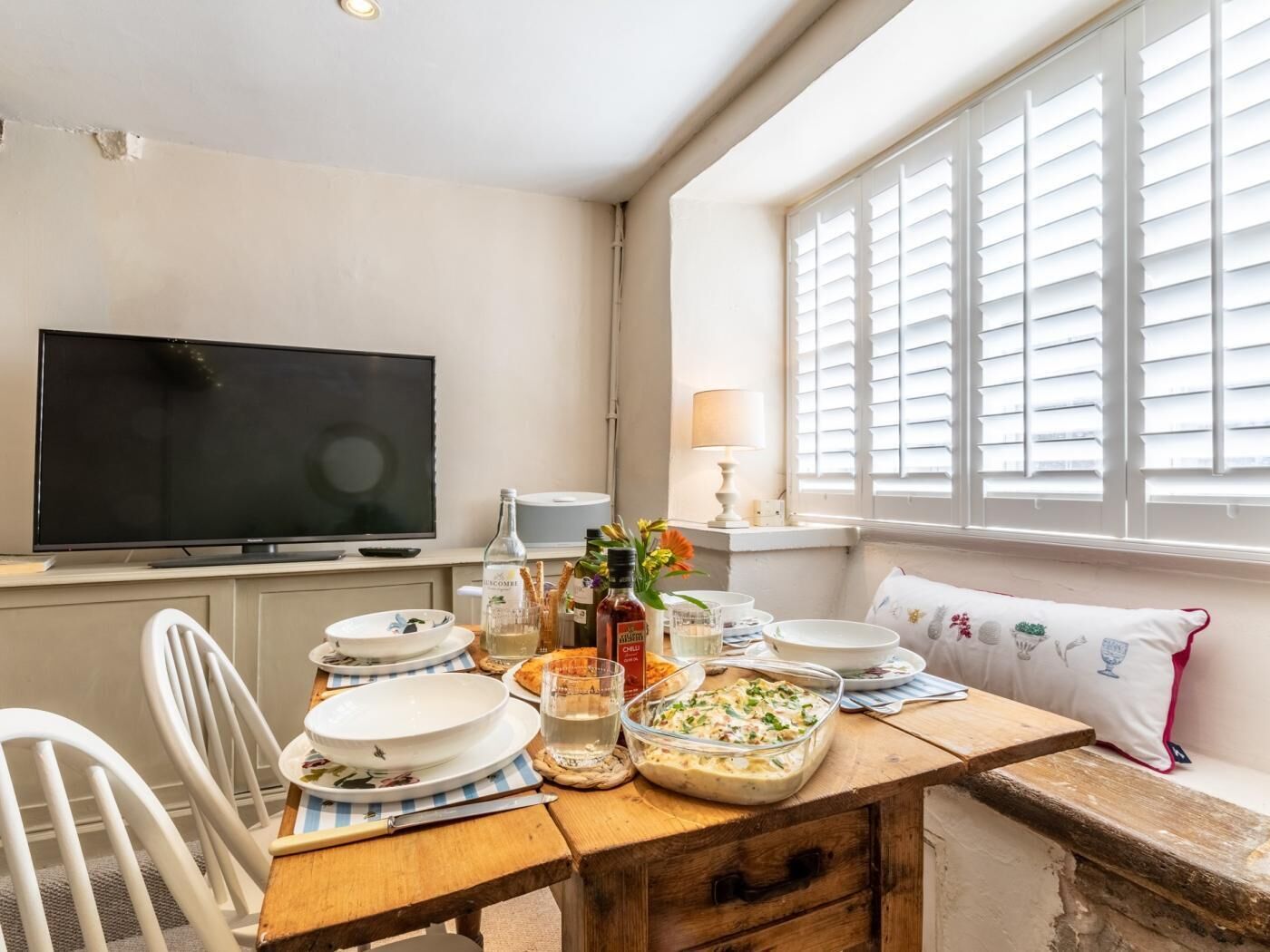 A dining area with a wooden table set for a meal, a television on the wall, and a window with shutters.