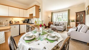 A modern kitchen with wooden cabinets, a dining area with a table set for four, and a living room with a sofa and a window. - 2 Bed in Beaworthy (oc-77555) (Black Torrington)