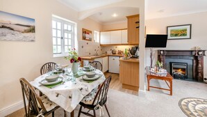 A dining area with a table set for four, a kitchen with wooden cabinets, a fireplace, and a television mounted on the wall. - 2 Bed in Beaworthy (oc-77555) (Black Torrington)