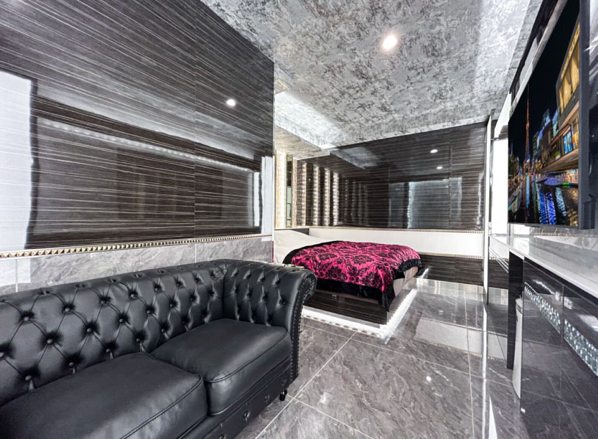 A modern hotel room with a black leather sofa, a bed with a patterned bedspread, and a large mirror reflecting the room.