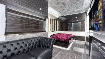 A modern hotel room with a black leather sofa, a bed with a patterned bedspread, and a large mirror reflecting the room.