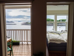 A room with a bed, a window with a view of the sea, and a balcony with a chair and plants. - Seaview apartment in Henningsvær (Vågan)