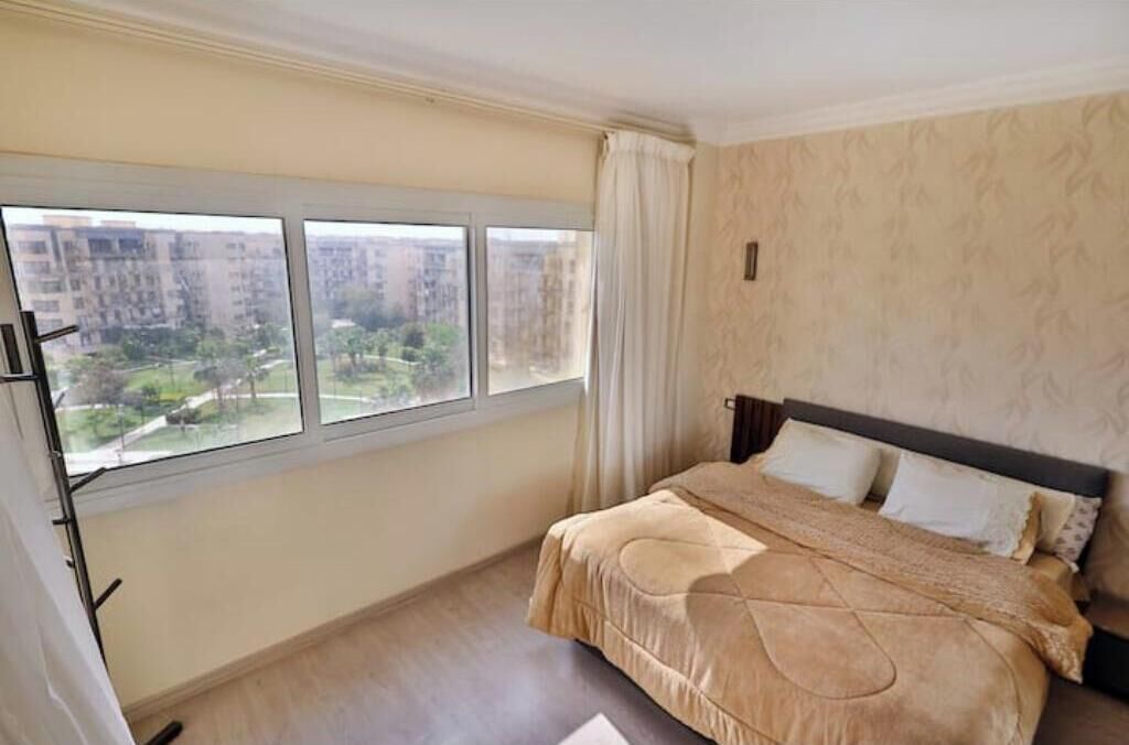 A bedroom with a large window, a bed with a beige bedspread, and a patterned wall.