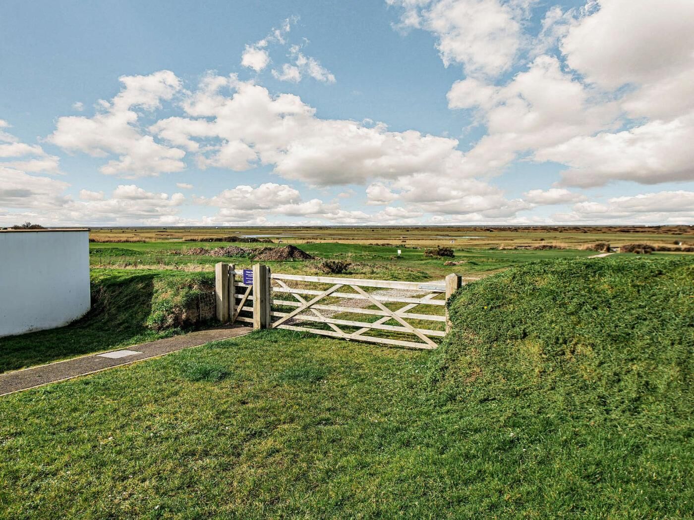 A rural landscape with a wooden gate, a white building, and open fields under a partly cloudy sky.