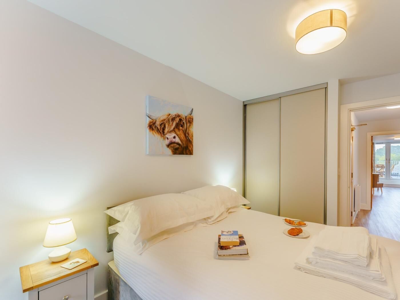 Ein Schlafzimmer mit einem Bett, einem Nachttisch mit Lampe, einem Gemälde und Blick ins Freie.