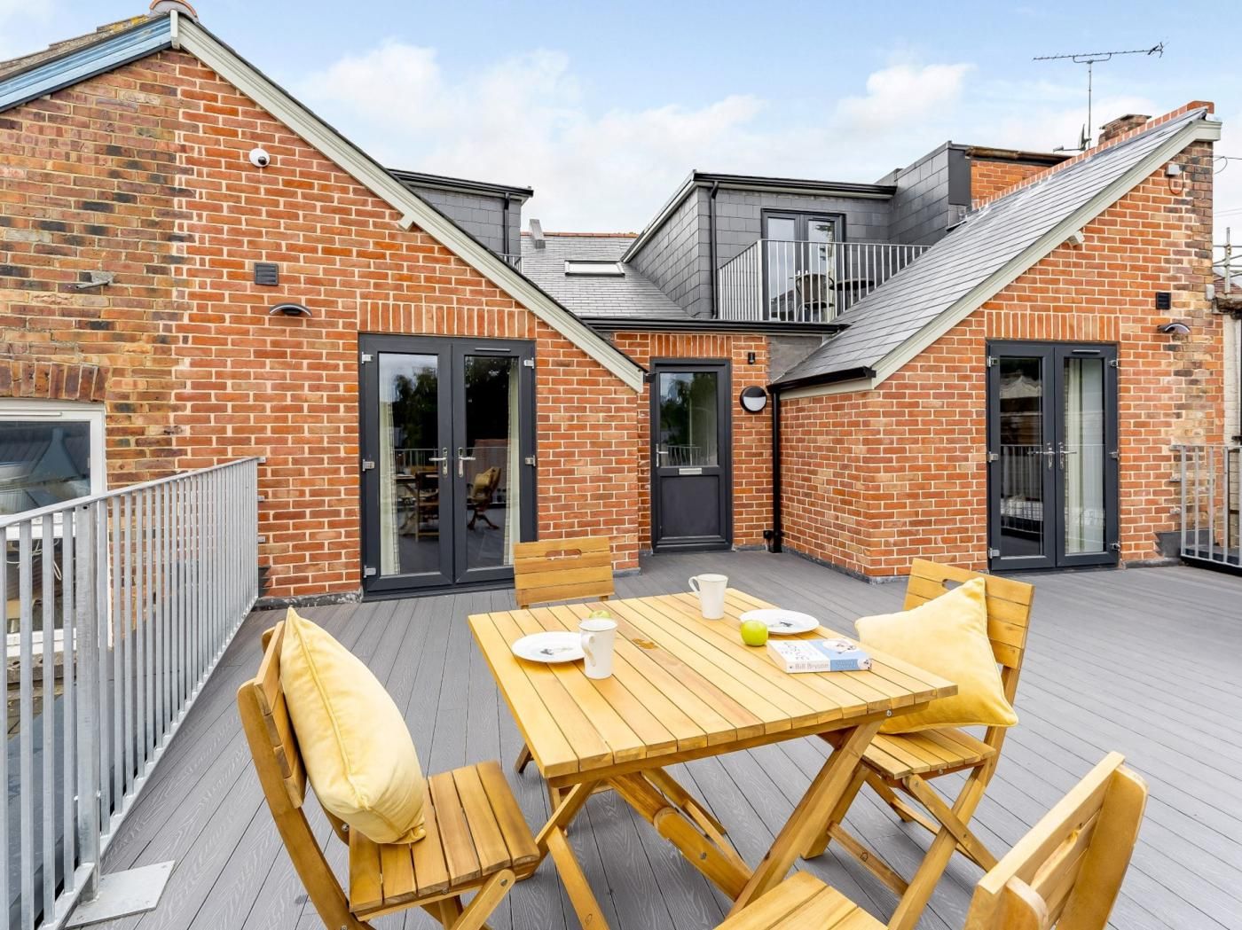 Une terrasse sur le toit avec une table et des chaises en bois, un bâtiment en briques avec une porte noire et un toit gris.