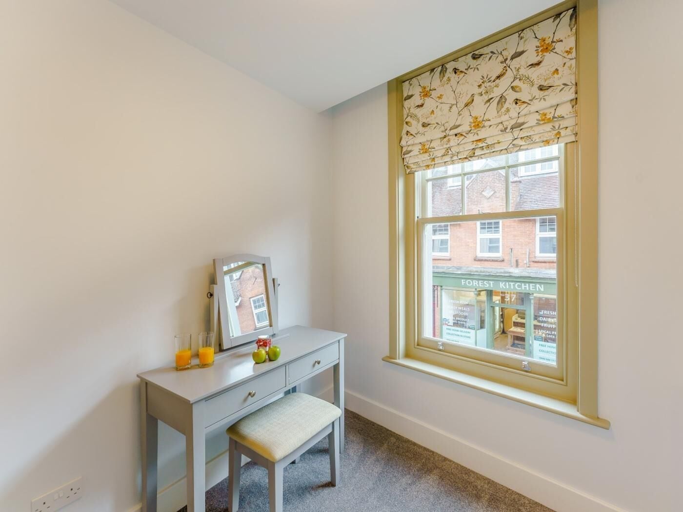 A corner room with a window, a desk with a mirror, a bench, and a view of a building with a sign.