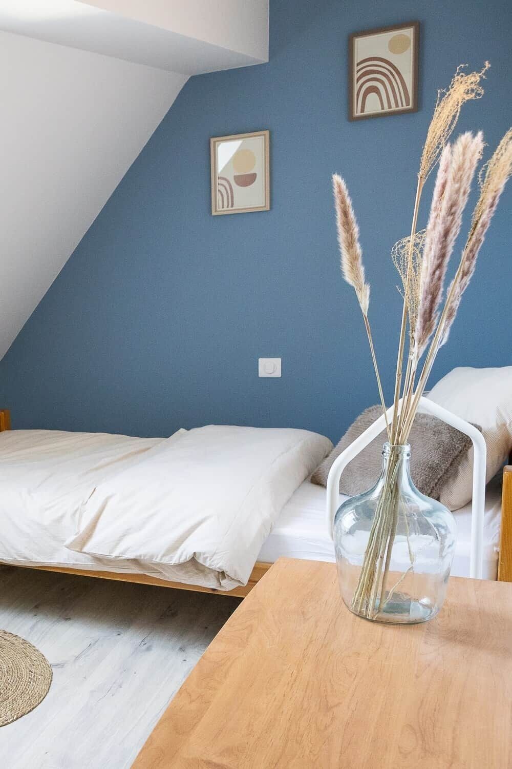 A bedroom with a bed, a wooden table, a vase with dried plants, and two wall-mounted pictures.