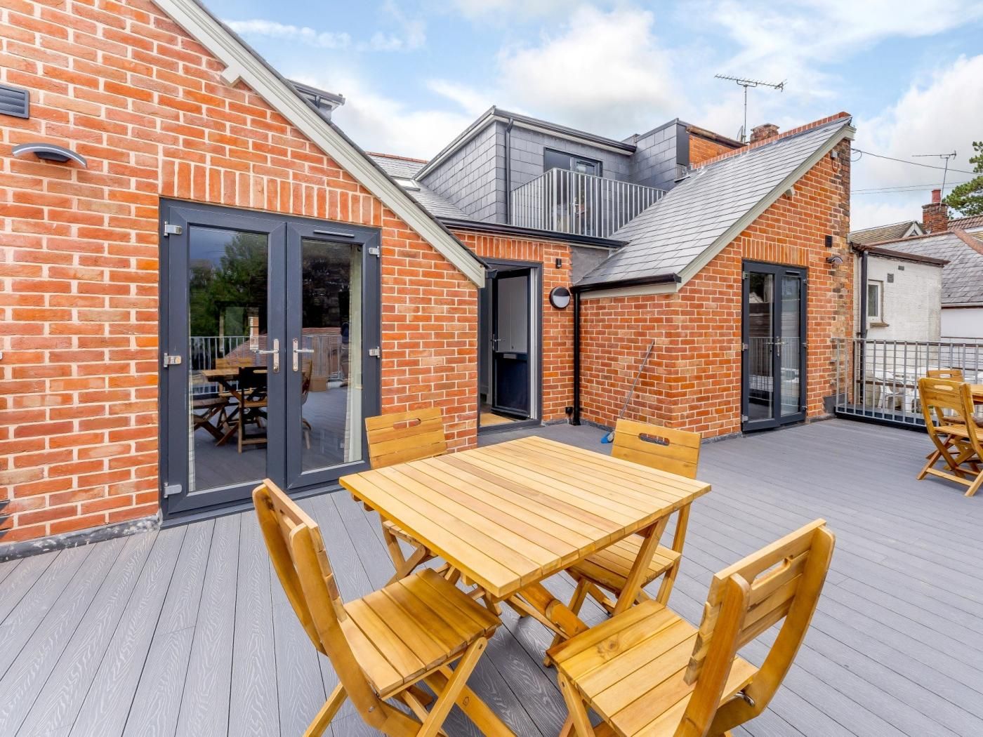 A wooden deck with a table and chairs, a glass door entrance, and a brick building with a sloped roof.