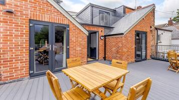 A wooden deck with a table and chairs, a glass door entrance, and a brick building with a sloped roof.