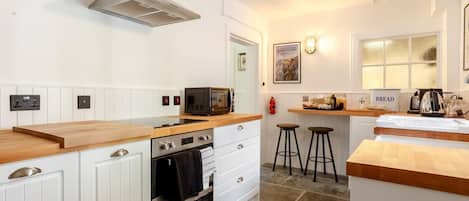 Una cocina moderna con armarios blancos, horno empotrado, microondas y encimera de madera. Hay dos taburetes y un cartel con la palabra 'pan' en la encimera.